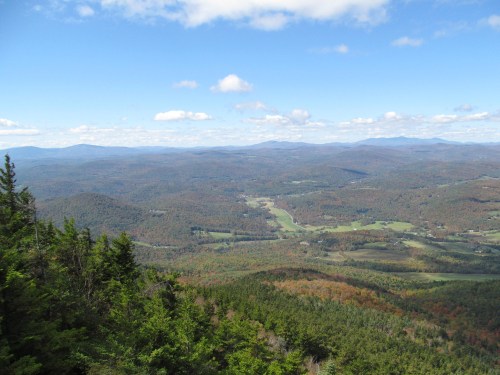 A panoramic view of rolling hills and valleys, with scattered trees and a blue sky dotted with clouds.