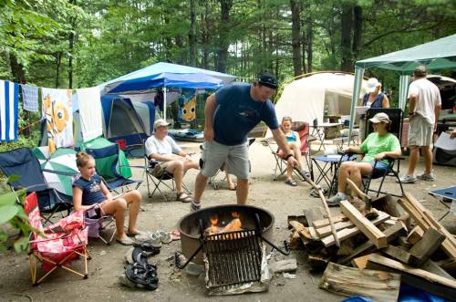 A family enjoys camping, gathered around a fire pit with tents and chairs in a wooded area.