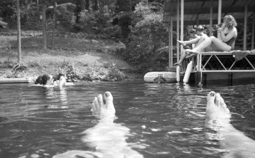 Black and white photo of feet in water, with people relaxing on a dock and swimming nearby.