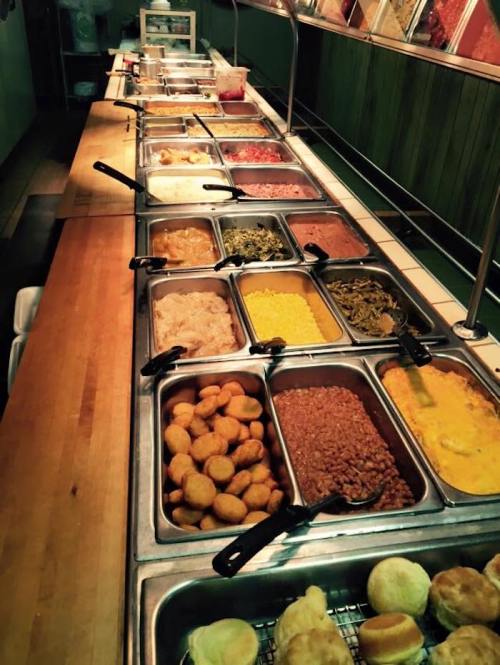 A buffet line featuring various dishes in metal trays, including sides like cornbread, greens, and casseroles.
