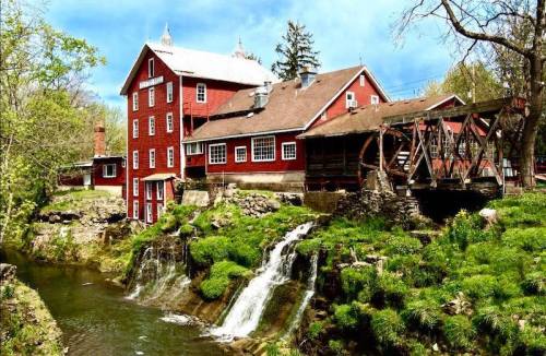 A red historic mill beside a stream with small waterfalls, surrounded by lush greenery and trees under a blue sky.