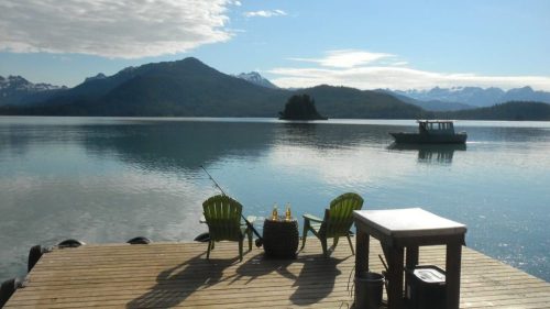 A serene lake view with two green chairs on a dock, mountains in the background, and a boat on the water.