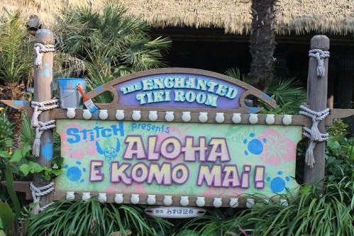 Sign for "The Enchanted Tiki Room" featuring Stitch, with the text "Aloha e komo mai!" surrounded by tropical decor.
