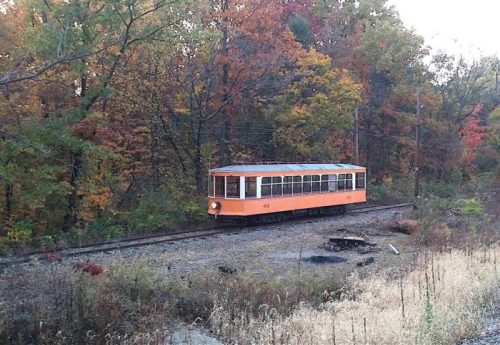 An orange vintage train car on a railway surrounded by colorful autumn trees.