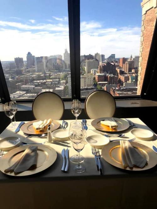 Elegant dining table set for two, overlooking a city skyline through large windows. Bright, sunny day.