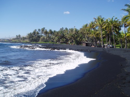 A scenic view of a black sand beach lined with palm trees under a clear blue sky and gentle waves.