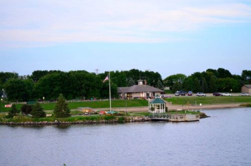 A serene lakeside view featuring a small dock, a flagpole, and a building surrounded by trees and grassy areas.
