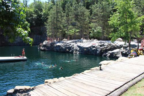 A scenic swimming area with people in the water, surrounded by rocky cliffs and lush trees, with a wooden walkway nearby.