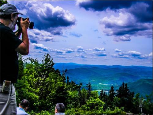 A photographer captures a scenic mountain view with clouds above, while others sit nearby enjoying the landscape.