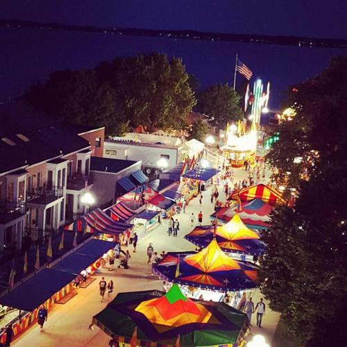 A vibrant nighttime scene of a fair with colorful tents, lights, and people enjoying the festivities by the water.
