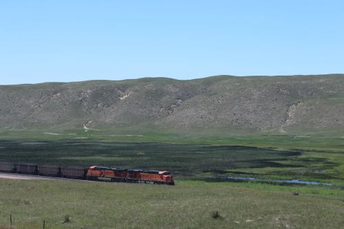A train travels through a green landscape with rolling hills and a small body of water under a clear blue sky.