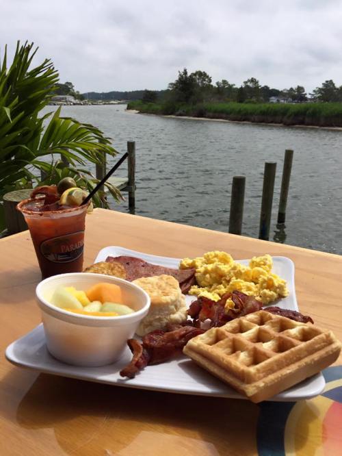 A plate of breakfast with bacon, eggs, biscuits, fruit, and a waffle, beside a drink, overlooking a scenic water view.