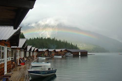 A serene lakeside scene with wooden cabins, boats, and a rainbow arching over misty mountains in the background.