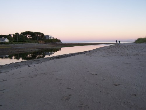 A serene beach at dusk, with a calm waterway and two people walking along the shore. Soft pastel sky in the background.