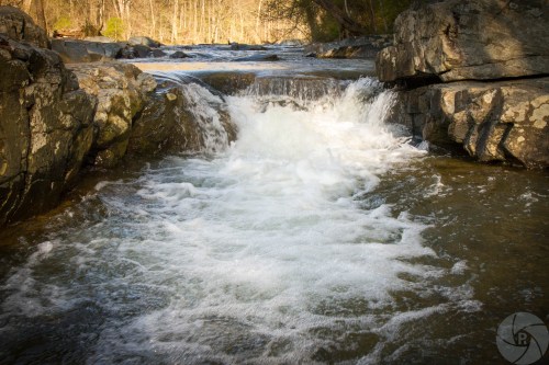 A small waterfall cascades over rocks into a serene river surrounded by trees.