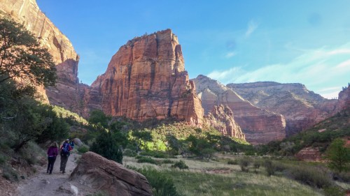 Two hikers walk along a trail in a canyon, surrounded by towering red rock formations and lush greenery.
