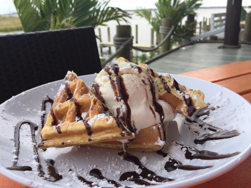 A plate of waffles topped with ice cream, chocolate sauce, and powdered sugar, set against a scenic outdoor backdrop.