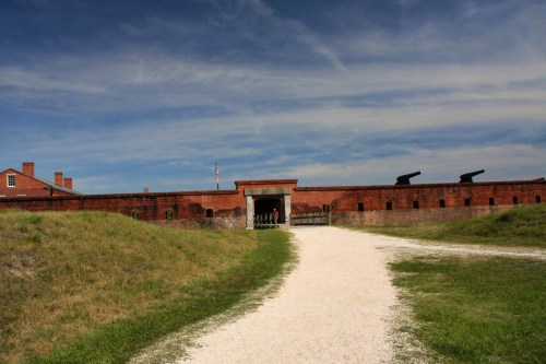 A pathway leads to a historic brick fort under a blue sky, with grassy areas on either side.