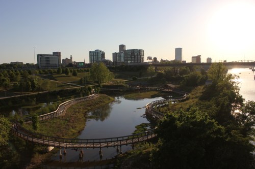 A winding boardwalk along a river with a city skyline in the background, illuminated by the setting sun.