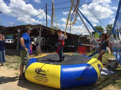A child bounces on a trampoline while harnessed, with adults nearby at a fair or outdoor event.