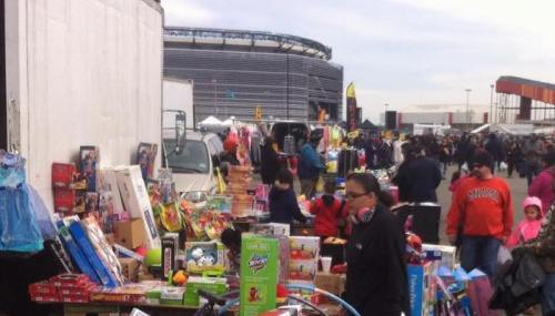 A bustling outdoor market with various toys and goods for sale, set against a stadium backdrop.