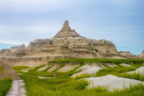 A rugged rock formation rises above green grass in a scenic landscape under a cloudy sky.