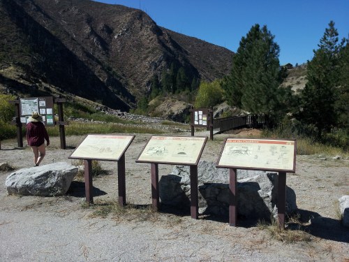 Information signs in a scenic outdoor area with mountains and trees in the background. A person walks nearby.