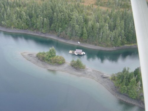 Aerial view of a serene river with a small dock and cabin surrounded by lush green trees and a sandy shoreline.