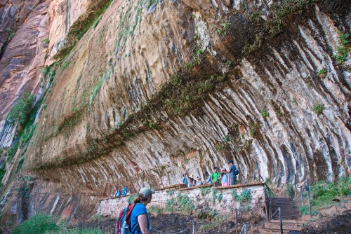 A hiker observes a large, textured rock wall with people exploring below in a scenic outdoor setting.