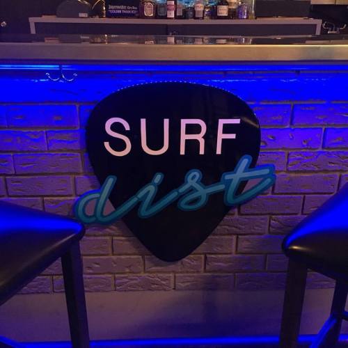 A neon sign reading "SURF dist" on a brick wall, with bar stools in the foreground.
