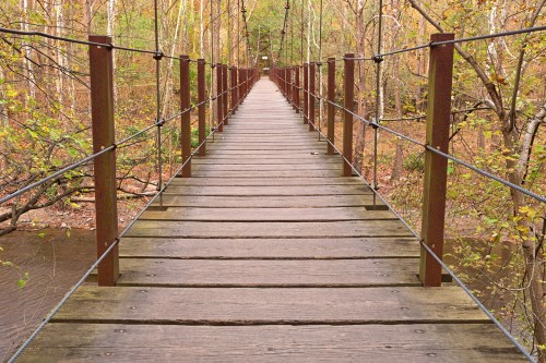 A wooden suspension bridge stretches over a calm river, surrounded by trees in autumn foliage.