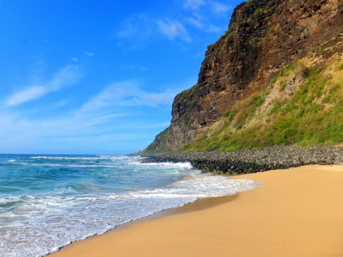 A sandy beach with gentle waves, bordered by a rocky cliff under a bright blue sky.