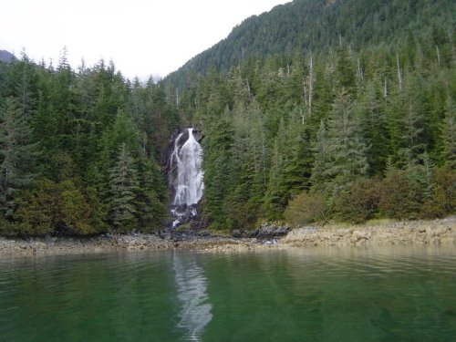A serene waterfall cascades down rocky cliffs, surrounded by lush green trees and reflecting in calm waters below.