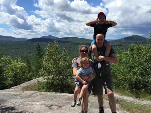 A family of four poses on a rocky outcrop with mountains and clouds in the background, enjoying a sunny day outdoors.