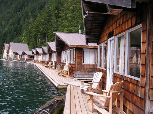 Wooden cabins line a waterfront, surrounded by lush green trees and calm water, with chairs on the porch.