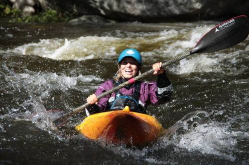 A smiling person in a kayak navigates through rushing water, wearing a blue helmet and a purple jacket.