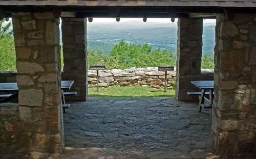 View from a stone shelter overlooking a green landscape and distant mountains, with picnic tables in the foreground.