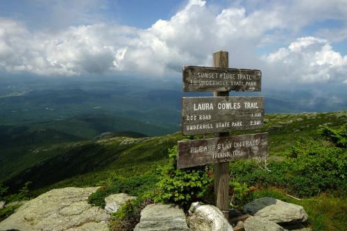 Signpost at a mountain trail intersection with scenic views and clouds in the background.