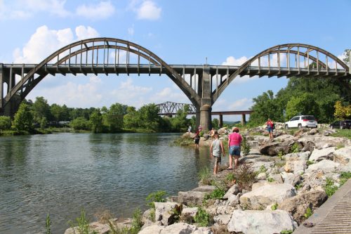 A scenic view of a bridge over a river, with people exploring the rocky shore and lush greenery in the background.