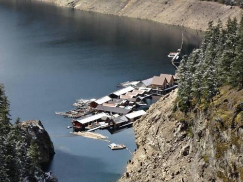 Aerial view of a snowy lakeside marina with wooden docks and cabins surrounded by rocky terrain and evergreen trees.