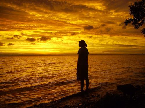 Silhouette of a person standing by the water at sunset, with vibrant orange and yellow clouds in the sky.
