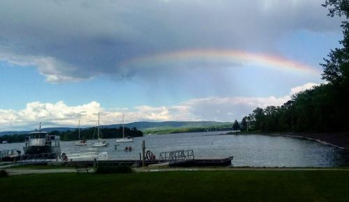 A serene lakeside view with a rainbow arching over the water, boats docked, and lush greenery in the background.