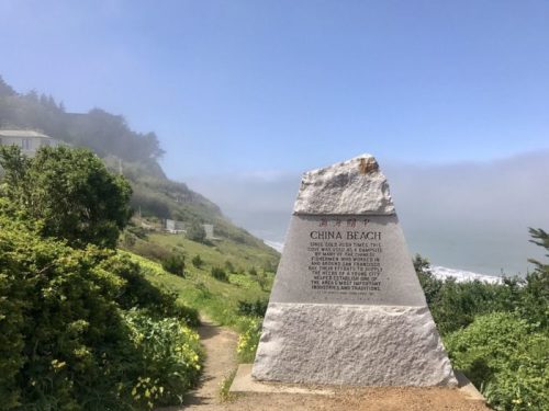 A stone monument at China Beach, surrounded by greenery and fog, with a view of the ocean in the background.