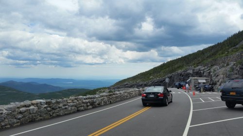 A winding road alongside a rocky hillside, with cars parked and a scenic view of mountains and cloudy skies in the background.