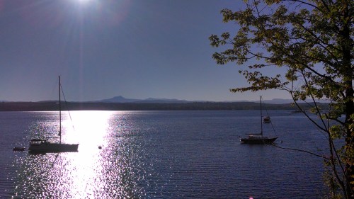 A serene lake scene with two sailboats, shimmering water, and distant mountains under a clear blue sky.