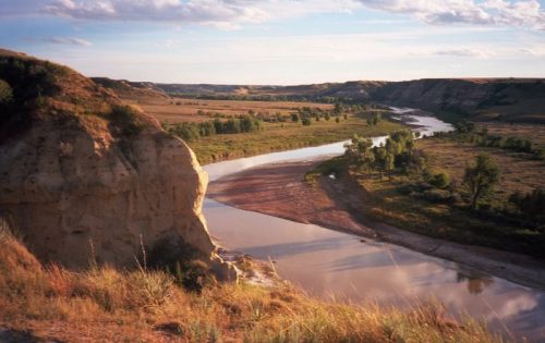 A winding river flows through a grassy landscape, flanked by cliffs and trees under a blue sky with scattered clouds.