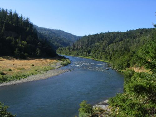 A serene river flows through a lush green valley, surrounded by hills and trees under a clear blue sky.