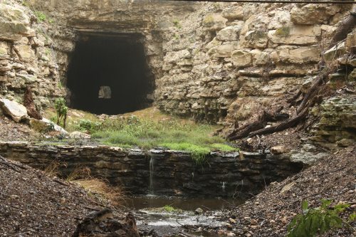 A rocky cave entrance surrounded by greenery and a small stream flowing in the foreground.
