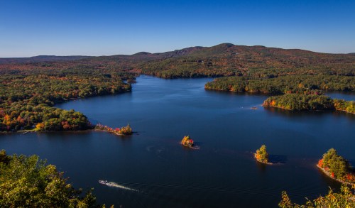 A scenic view of a tranquil lake surrounded by colorful autumn foliage and distant mountains under a clear blue sky.
