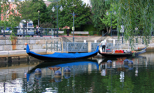 Two gondolas docked by a serene waterway, surrounded by greenery and a peaceful urban setting.
